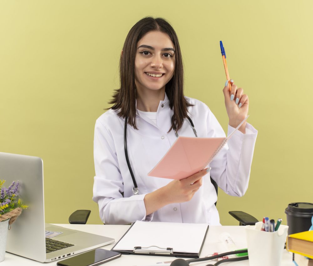 young woman doctor in white coat with stethoscope around her neck holding notebook and pen looking at camera wirh smile on face sitting at the table with laptop over light background